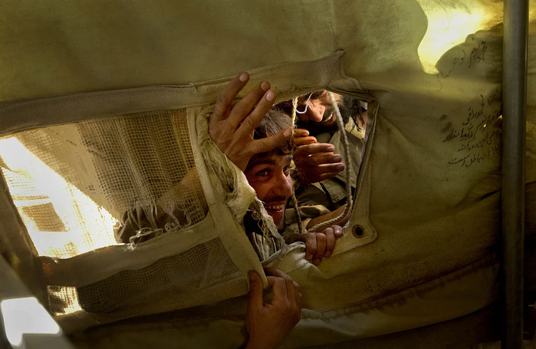 Men and boys peer through a torn opening in a tent, with hands gripping the fabric.