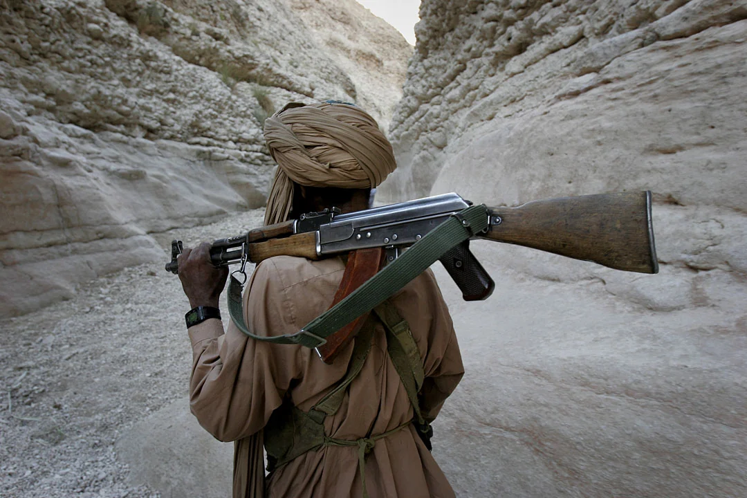 A Baloch fighter seen from behind carrying a weapon through a pale, narrow canyon.