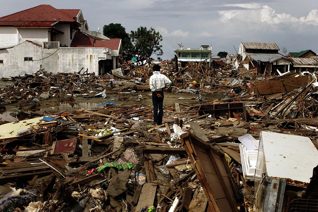 A person walks through extensive rubble between damaged buildings after a tsunami.
