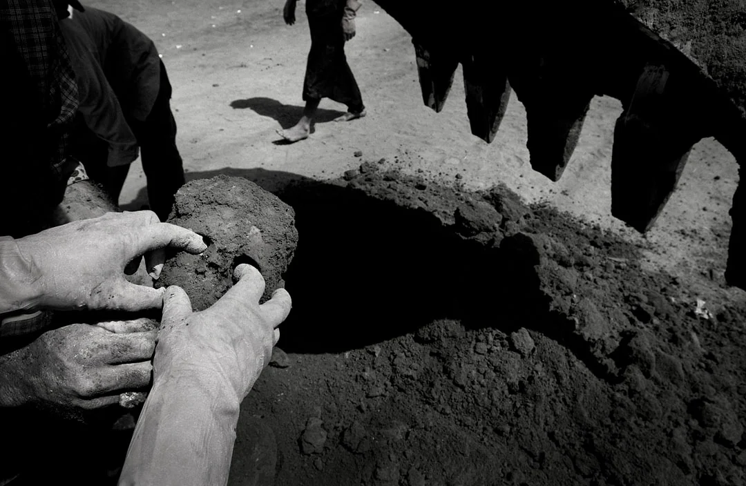 Hands hold a skull beside an open grave, with people standing nearby, photographed in black and white.