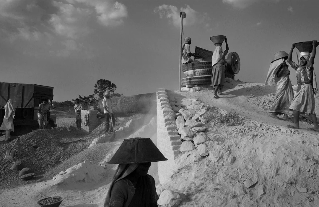 Workers carrying materials through a stone-crushing site in a black-and-white photograph.