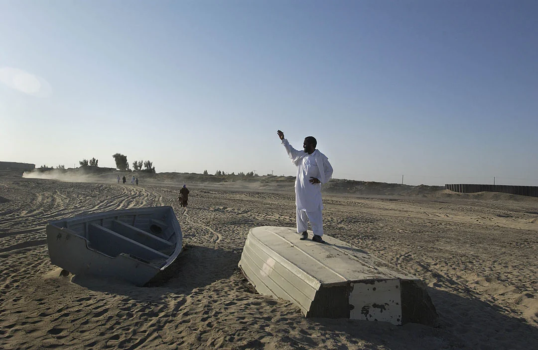 A man stands on a stranded boat in a dry riverbed, gesturing across the sand.