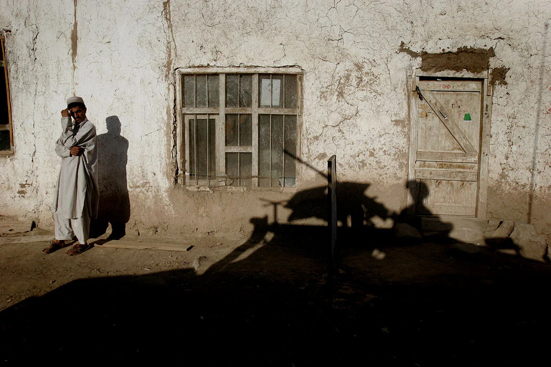 A man stands against a sunlit wall above the dark shadow of a military vehicle.