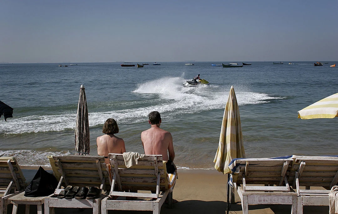 Two people in beach chairs watch a jet ski beyond folded umbrellas at the water's edge.