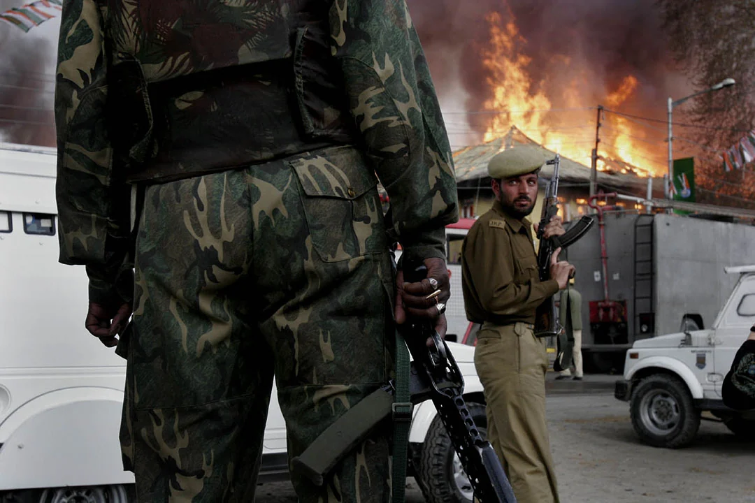 Armed soldiers stand in the foreground as flames and smoke rise from a building behind them.