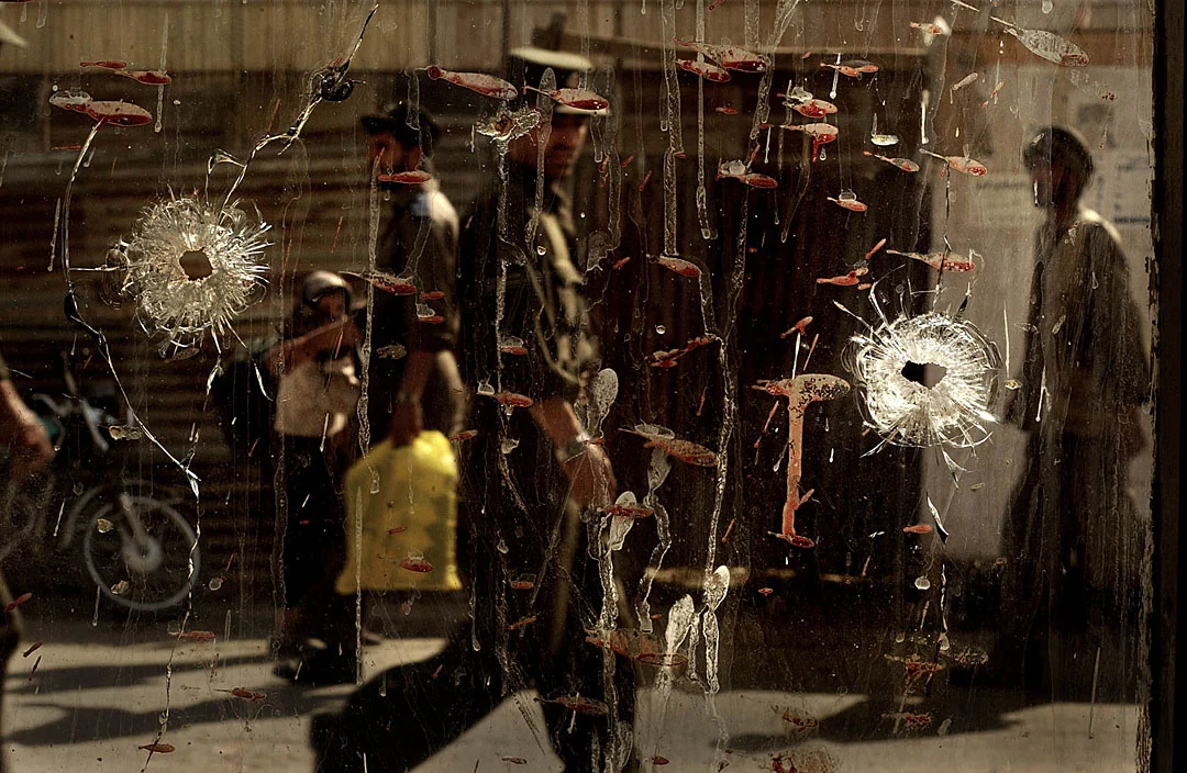 People on a street seen through shattered glass marked by dried blood after a bombing.
