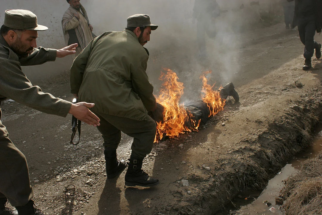 Police officers pull a fallen colleague away from flames after a bombing, with smoke rising around them.