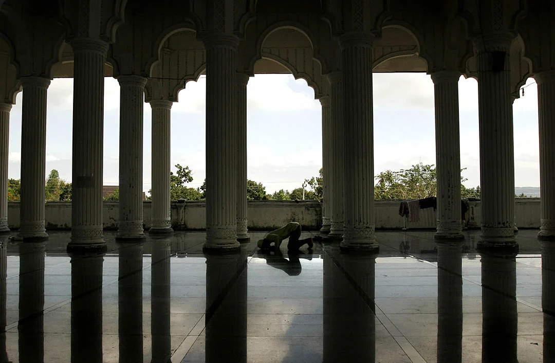 A solitary person praying between rows of dark columns in a mosque.