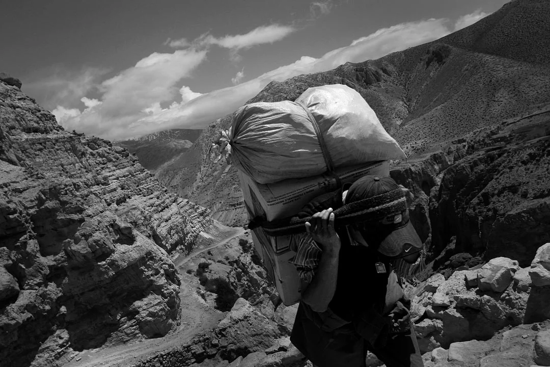 A porter carrying a large load along a rocky mountain path, in black and white.