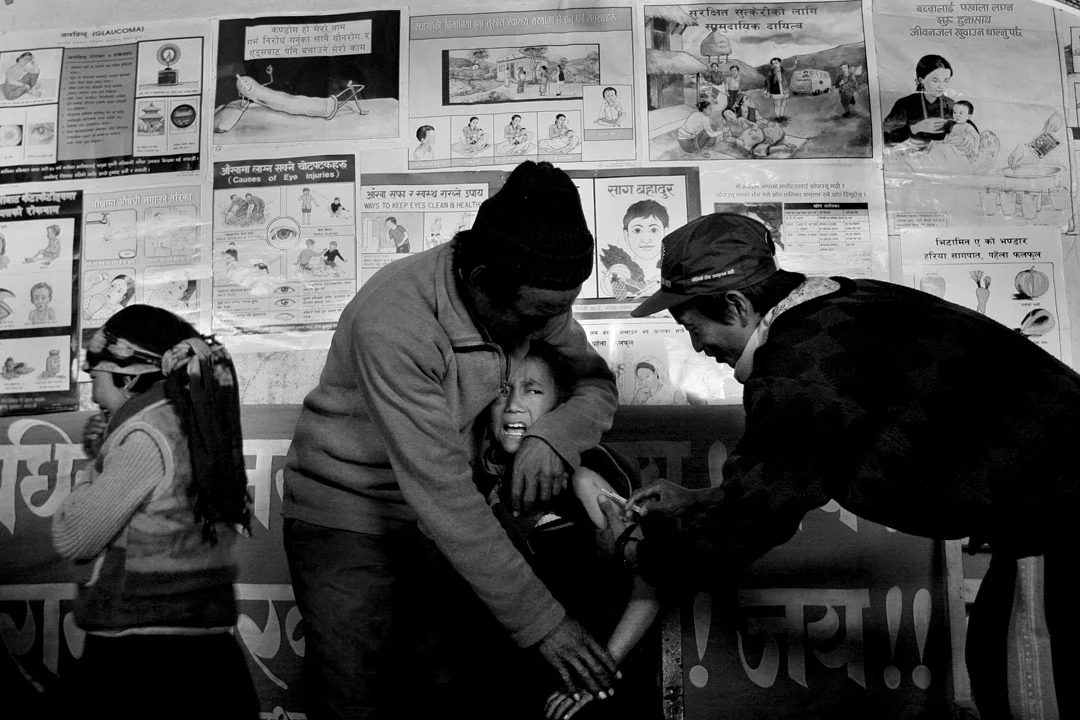A child receiving a vaccination in a health post, surrounded by people and posters.