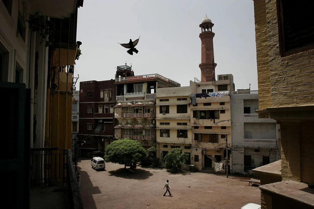 A bird flies above a courtyard enclosed by old buildings, with a minaret in the distance.