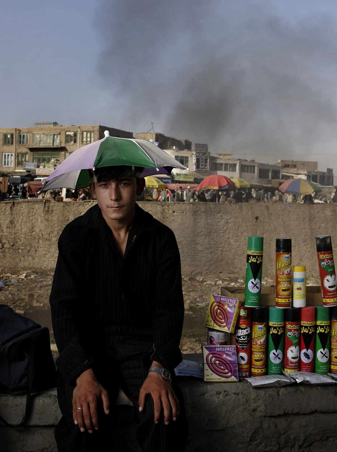 An insecticide seller wearing a green hat sits beside rows of bottles, with smoke behind him.