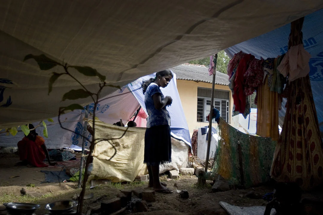 A woman stands beneath the fabric roof of a temporary shelter, surrounded by household belongings.